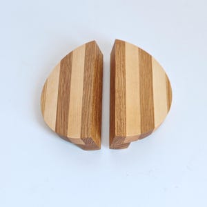 May include: Two semi-circular wooden drawer pulls featuring alternating vertical stripes of light and medium-toned wood grain. Each handle has a flat top edge and a curved outer side, showcasing the natural texture and colour variations of the wood.