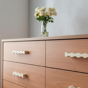 May include: A light brown wooden dresser featuring four drawers with horizontal, wavy-shaped off-white drawer pulls. The dresser has a smooth, flat top surface and a clean, rectangular structure with visible wood grain patterns on the drawer fronts.