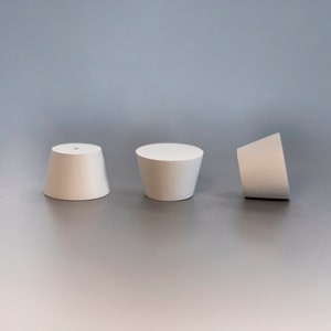 May include: Three white, truncated cone-shaped stoppers are arranged in a row. Each stopper features a smooth, matte surface and a flat circular top. The leftmost stopper has a small, centered hole, while the others have solid, flat tops.