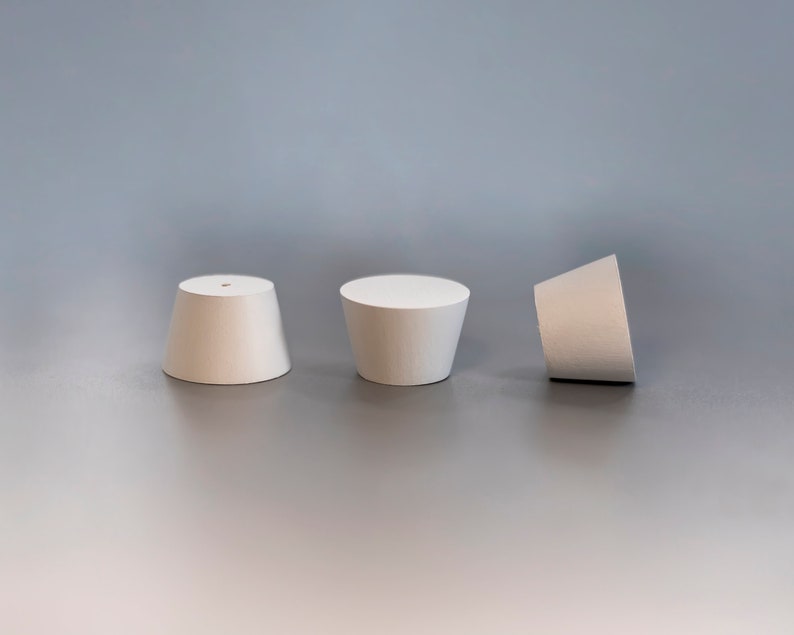 May include: Three white, truncated cone-shaped stoppers are arranged in a row. Each stopper features a smooth, matte surface and a flat circular top. The leftmost stopper has a small, centered hole, while the others have solid, flat tops.