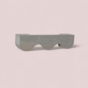 May include: A gray wooden object with a wavy design. The object has a rectangular shape with three curved cutouts along the bottom edge. The wood grain is visible, and the object is set against a light pink background.