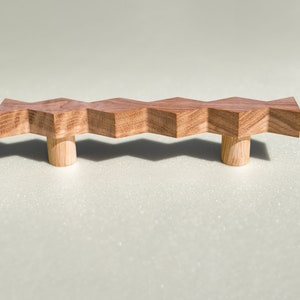 Puede incluir: Un tirador de cajón de madera con un diseño en zigzag y dos pomos redondos. El tirador está hecho de madera marrón claro y tiene un acabado natural.
