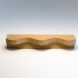 May include: A wooden drawer pull with a wavy design. The pull is made of light brown wood and has a smooth finish.