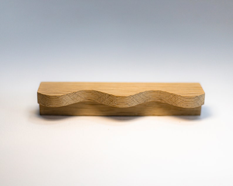 May include: A wooden drawer pull with a wavy design. The pull is made of light brown wood and has a smooth finish.