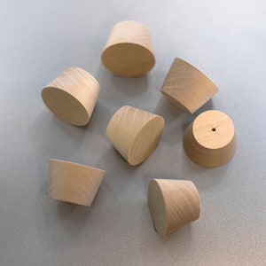 May include: A collection of six light-colored, unfinished wooden knobs with a tapered, conical shape. Each knob features a smooth, natural wood grain texture and a flat circular top surface. One knob displays a small, centered hole on its top.
