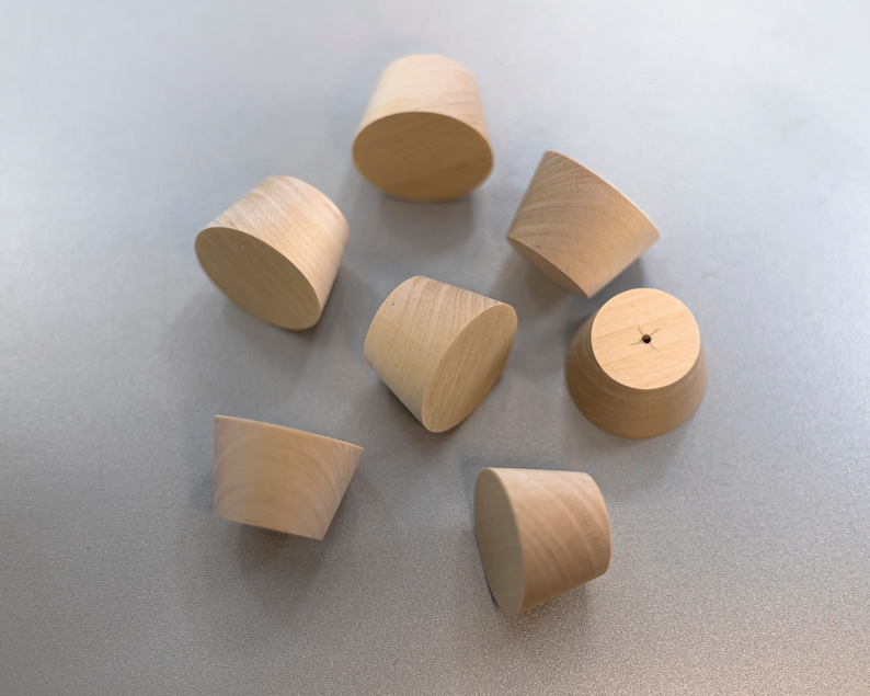 May include: A collection of six light-colored, unfinished wooden knobs with a tapered, conical shape. Each knob features a smooth, natural wood grain texture and a flat circular top surface. One knob displays a small, centered hole on its top.