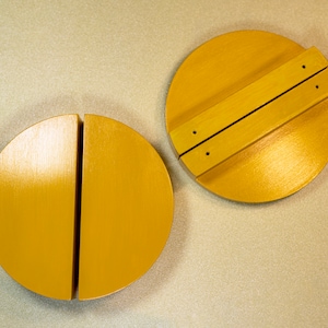 Op de afbeelding: Twee gele houten deksels, een met een scharnierend handvat, de andere in tweeën gesplitst.