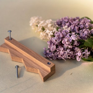 Puede incluir: Un tirador de cajón de madera con un diseño en zigzag y dos tornillos plateados. El tirador está hecho de madera marrón claro y tiene un acabado natural. El tirador está sobre una superficie blanca con un ramo de flores de lila púrpura y blanco.