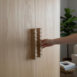 May include: A light-colored wooden cabinet door featuring a vertical handle with a distinct wavy, rippled edge design. The handle is crafted from a similar light-toned wood, showcasing a natural grain pattern that matches the cabinet surface.