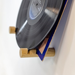 May include: A close-up of a vinyl record holder made of wood. The holder is attached to a white wall and has two wooden pegs that hold the record in place. The record is black and has a blue and pink album cover.