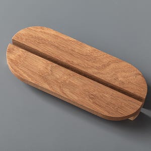 Puede incluir: Un soporte de exhibición de madera de forma ovalada con un acabado de grano de madera marrón claro natural y una ranura recta y estrecha cortada a través del centro de la superficie superior, diseñado para sostener objetos finos.