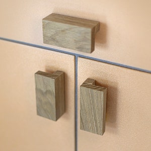 May include: Three wooden cabinet pulls, two are rectangular and one is a longer rectangle. The pulls are a light brown colour and have a wood grain pattern.