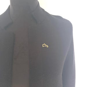 May include: A black blazer with a gold-colored pin in the shape of a map outline. The blazer is displayed on a mannequin against a white background. The pin is small and centered on the blazer.