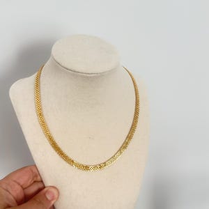 May include: A gold-colored necklace with a woven chain design. The necklace is displayed on a cream-colored bust against a white background. The chain is approximately 16 inches long.
