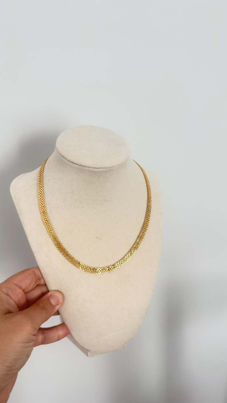 May include: A gold-colored necklace with a woven chain design. The necklace is displayed on a cream-colored bust against a white background. The chain is approximately 16 inches long.