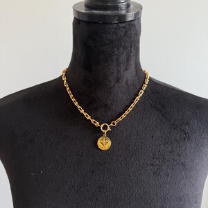 May include: A gold-colored necklace with a coin pendant. The necklace features a chunky chain design and a circular clasp. The coin pendant has a detailed embossed design. The necklace is displayed on a black mannequin.