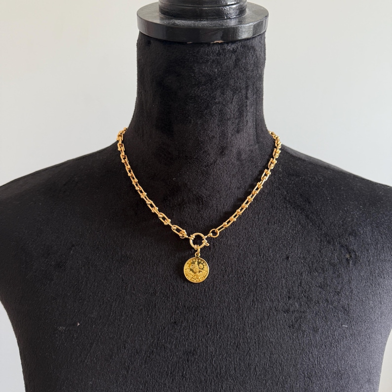 May include: A gold-colored necklace with a coin pendant. The necklace features a chunky chain design and a circular clasp. The coin pendant has a detailed embossed design. The necklace is displayed on a black mannequin.