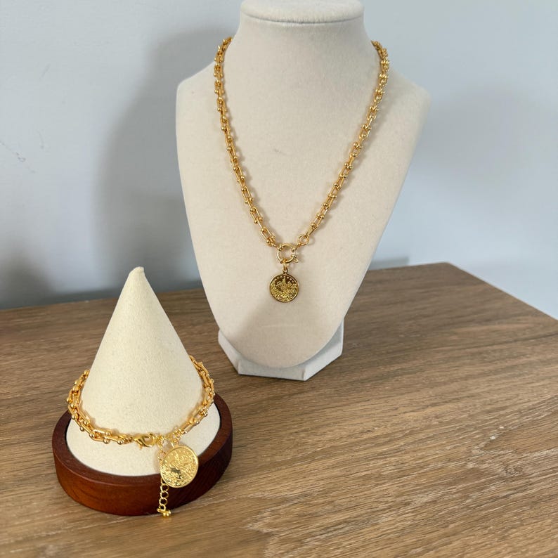 May include: A gold-colored necklace and bracelet set. The necklace is displayed on a white bust, and the bracelet is on a small wooden stand. Both pieces feature a chain design with a coin pendant.
