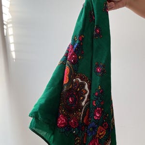 May include: A green scarf with a floral design. The scarf has a dark green base, adorned with a detailed pattern of red, pink, blue, and yellow flowers. The scarf is held up against a white backdrop.