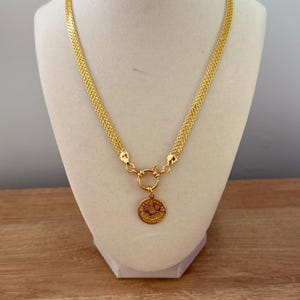 May include: A gold-colored necklace with a circular pendant. The necklace has a woven chain design and a round clasp. The pendant features a detailed design. The necklace is displayed on a white mannequin bust.