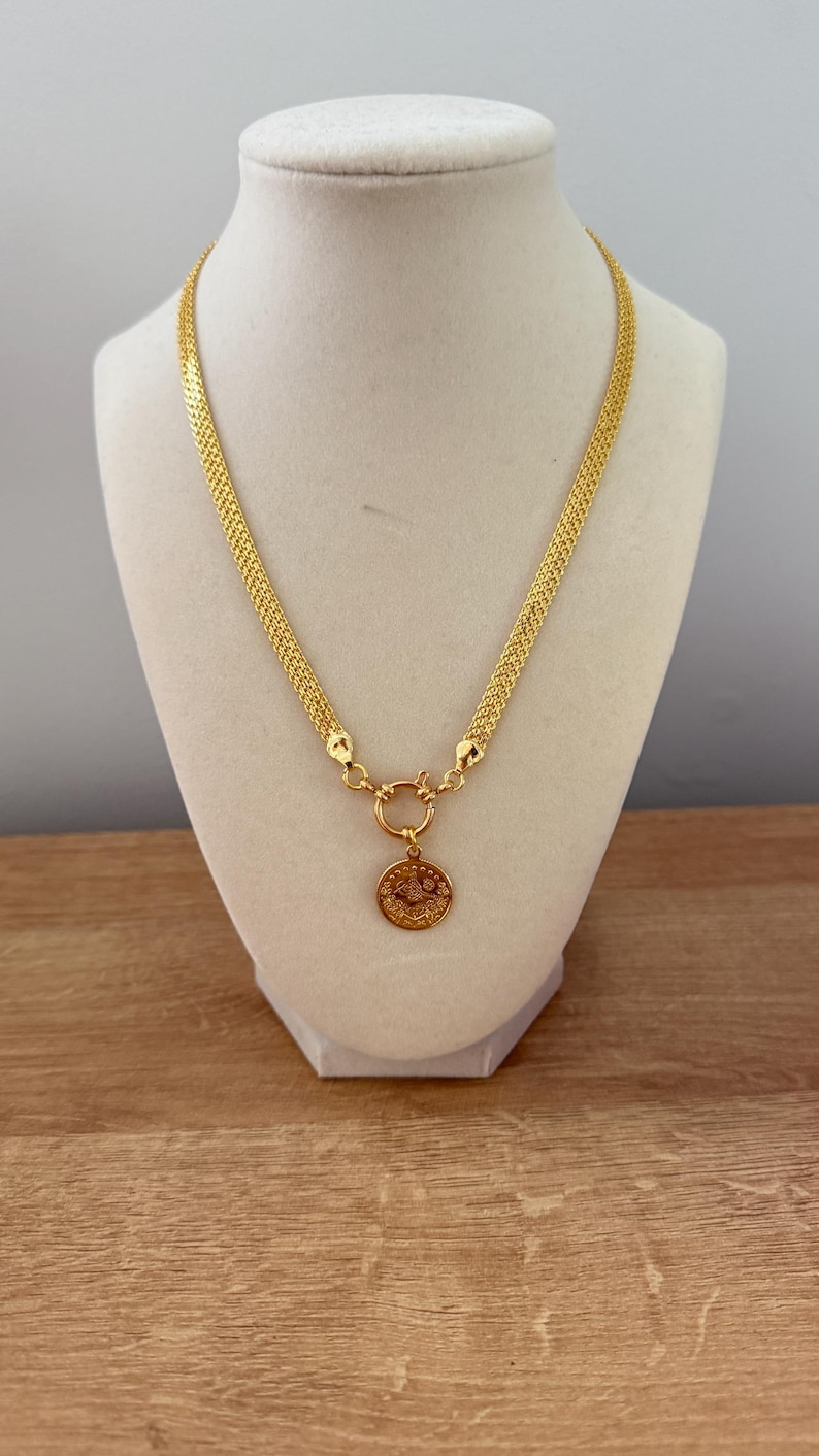 May include: A gold-colored necklace with a circular pendant. The necklace has a woven chain design and a round clasp. The pendant features a detailed design. The necklace is displayed on a white mannequin bust.