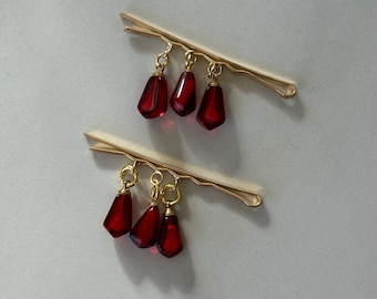 Pode incluir: Dois grampos de cabelo dourados com pingentes de vidro vermelho em forma de lágrima. Cada grampo tem três pingentes.