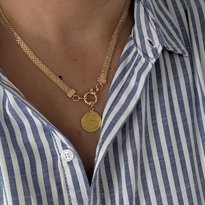 May include: Gold-toned necklace with a coin pendant. The necklace features a woven chain design and a circular coin pendant with embossed details. The person is wearing a blue and white striped button-down shirt.