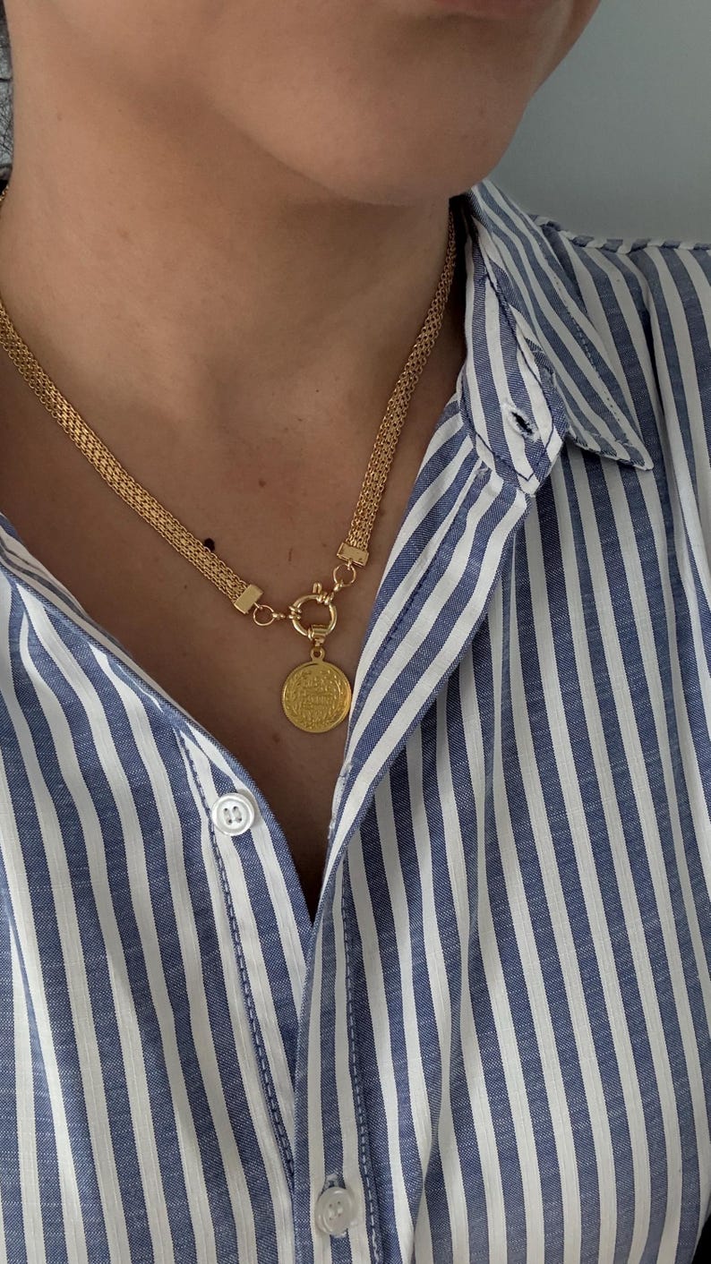 May include: Gold-toned necklace with a coin pendant. The necklace features a woven chain design and a circular coin pendant with embossed details. The person is wearing a blue and white striped button-down shirt.