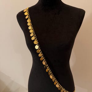 May include: A gold body chain with coin pendants. The chain is displayed on a black mannequin. The round coins are evenly spaced along the chain, creating a decorative accessory. The chain is designed to be worn across the body.