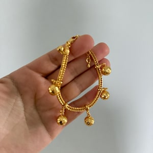 Op de afbeelding: Een goudkleurige armband met een kettingontwerp en bungelende balbedels. De armband wordt in een hand gehouden tegen een witte achtergrond. De ketting is gemaakt van in elkaar verweven schakels en de bedels zijn rond met een textuur.