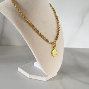 May include: Gold-toned necklace with a coin pendant. The necklace features a chain of small, round discs and a circular pendant with a detailed design. The jewelry is displayed on a white mannequin bust against a neutral background.