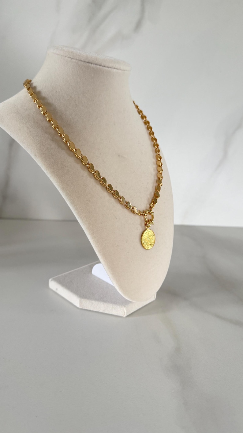 May include: Gold-toned necklace with a coin pendant. The necklace features a chain of small, round discs and a circular pendant with a detailed design. The jewelry is displayed on a white mannequin bust against a neutral background.