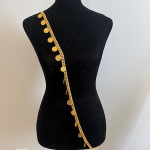 Puede incluir: Un accesorio corporal de cadena en tono dorado que presenta una serie de pequeños dijes circulares similares a monedas, unidos a intervalos regulares a lo largo de la cadena. El accesorio está drapeado diagonalmente sobre un torso de maniquí negro.