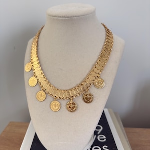 May include: A gold-colored necklace with coin pendants displayed on a mannequin. The necklace features a series of connected, curved segments, with several circular coin-shaped pendants hanging below. The coins have detailed designs. The necklace is presented on a cream-colored bust.