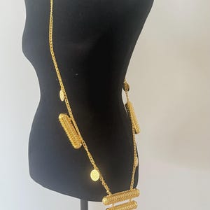 Puede incluir: Un collar largo de cadena en tono dorado con múltiples colgantes cilíndricos de textura de malla y pequeños dijes circulares similares a monedas. El diseño incluye una disposición escalonada de tres cilindros horizontales en la base.