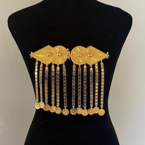 Puede incluir: Un broche de metal dorado con dos placas simétricas en forma de diamante con centros circulares y patrones radiales texturizados. Diez cadenas verticales cuelgan de las placas, cada una terminada en un pequeño disco de metal dorado.