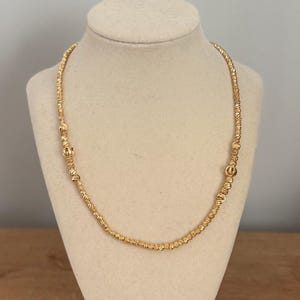 May include: A gold-colored necklace displayed on a mannequin. The necklace features a textured, beaded design with small, round accents. The necklace is draped in a gentle curve, showcasing its length and style.