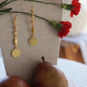 May include: A pair of gold dangle earrings with a coin charm. The earrings feature a chain with small beads and a decorative knot design. The earrings are displayed on a mannequin with red carnations and pears.
