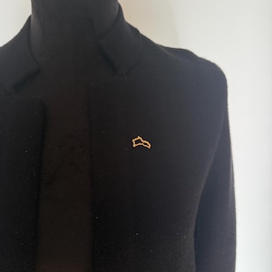 May include: A black blazer with a gold-colored pin in the shape of a stylized outline. The blazer is displayed on a mannequin, showcasing its tailored fit and textured fabric. The pin adds a touch of subtle elegance.