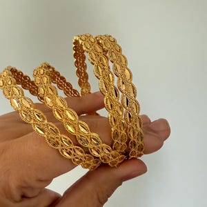 May include: Several gold-colored bangles with an intricate oval pattern are held in a hand. The bangles have a textured surface and a shiny finish. The jewelry is displayed against a plain white background.
