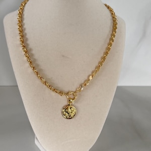 May include: Gold-toned necklace with a coin pendant. The necklace features a chain with a mix of rope and flat link designs. The coin pendant has a detailed embossed design. The necklace is displayed on a cream-colored bust.
