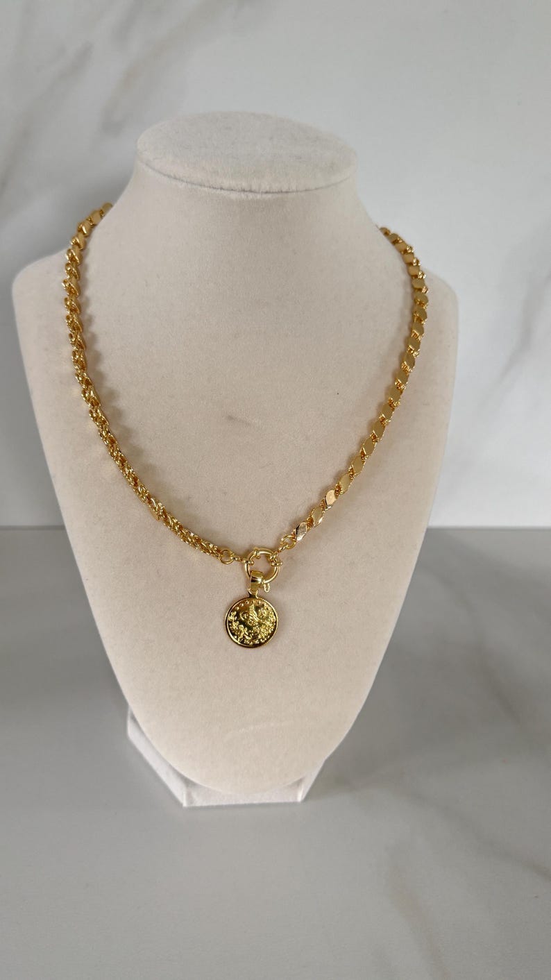 May include: Gold-toned necklace with a coin pendant. The necklace features a chain with a mix of rope and flat link designs. The coin pendant has a detailed embossed design. The necklace is displayed on a cream-colored bust.