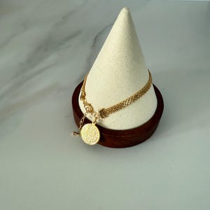 May include: A gold-colored bracelet with a delicate chain design and a round pendant. The bracelet is displayed on a white cone-shaped stand with a dark brown wooden base. The pendant has a textured surface.