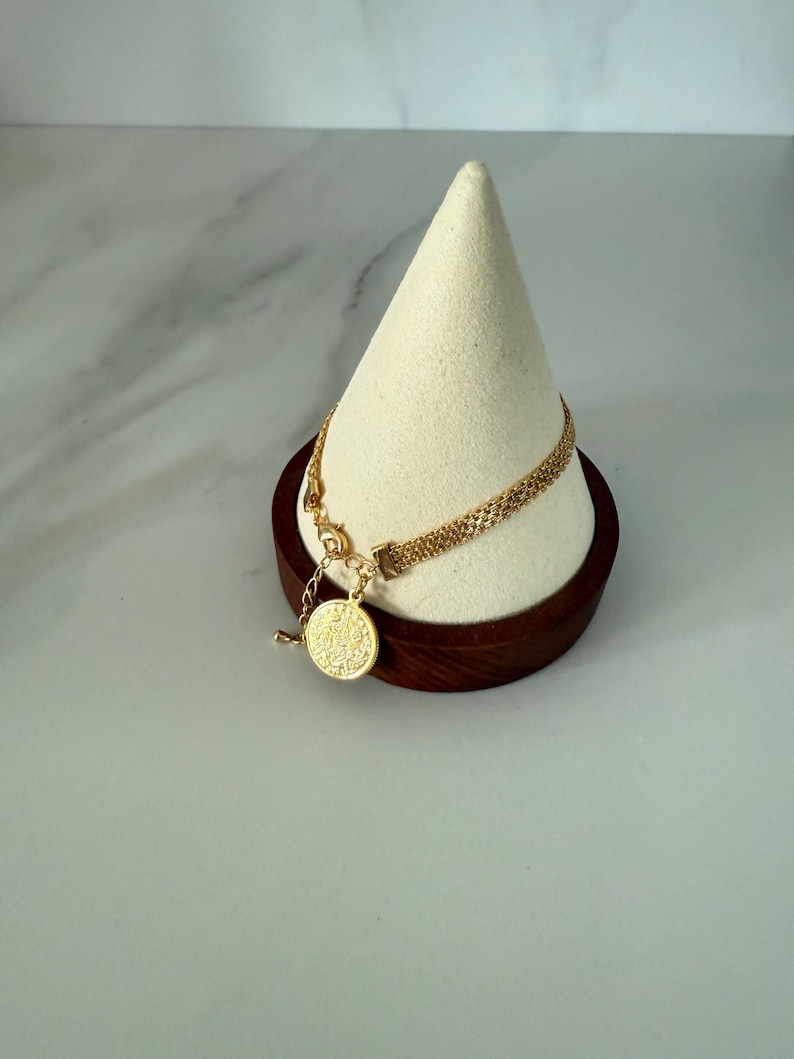 May include: A gold-colored bracelet with a delicate chain design and a round pendant. The bracelet is displayed on a white cone-shaped stand with a dark brown wooden base. The pendant has a textured surface.