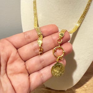 May include: A gold-colored necklace with a coin pendant. The necklace features a woven chain design and a circular clasp. The coin pendant has an embossed design. The necklace is displayed on a mannequin.