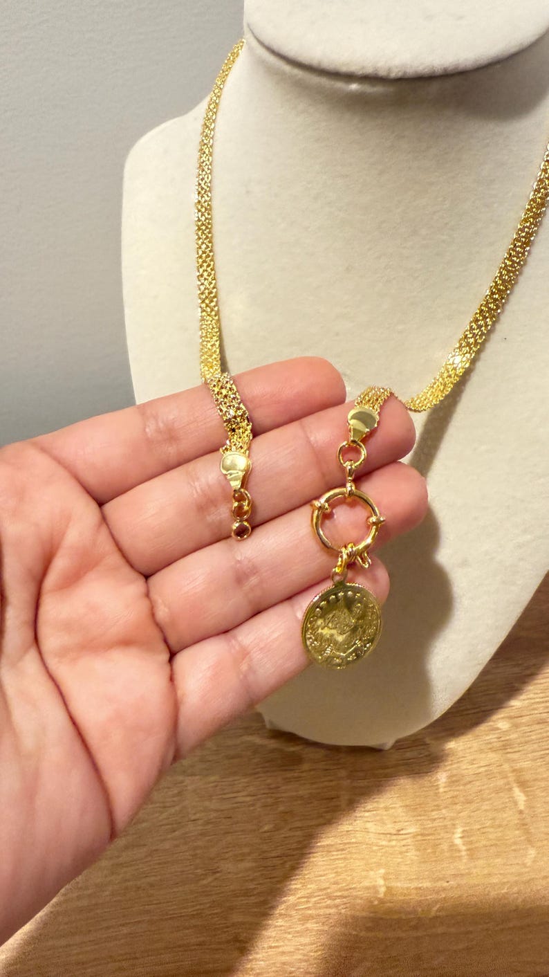 May include: A gold-colored necklace with a coin pendant. The necklace features a woven chain design and a circular clasp. The coin pendant has an embossed design. The necklace is displayed on a mannequin.