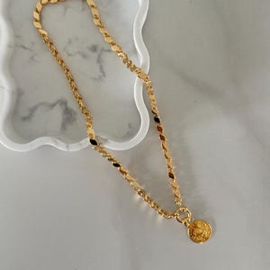 May include: A gold-colored necklace with a coin pendant rests on a white marble tray. The necklace features a variety of chain styles, including a flat, oval link design and a twisted rope chain. The coin pendant has a detailed embossed design.