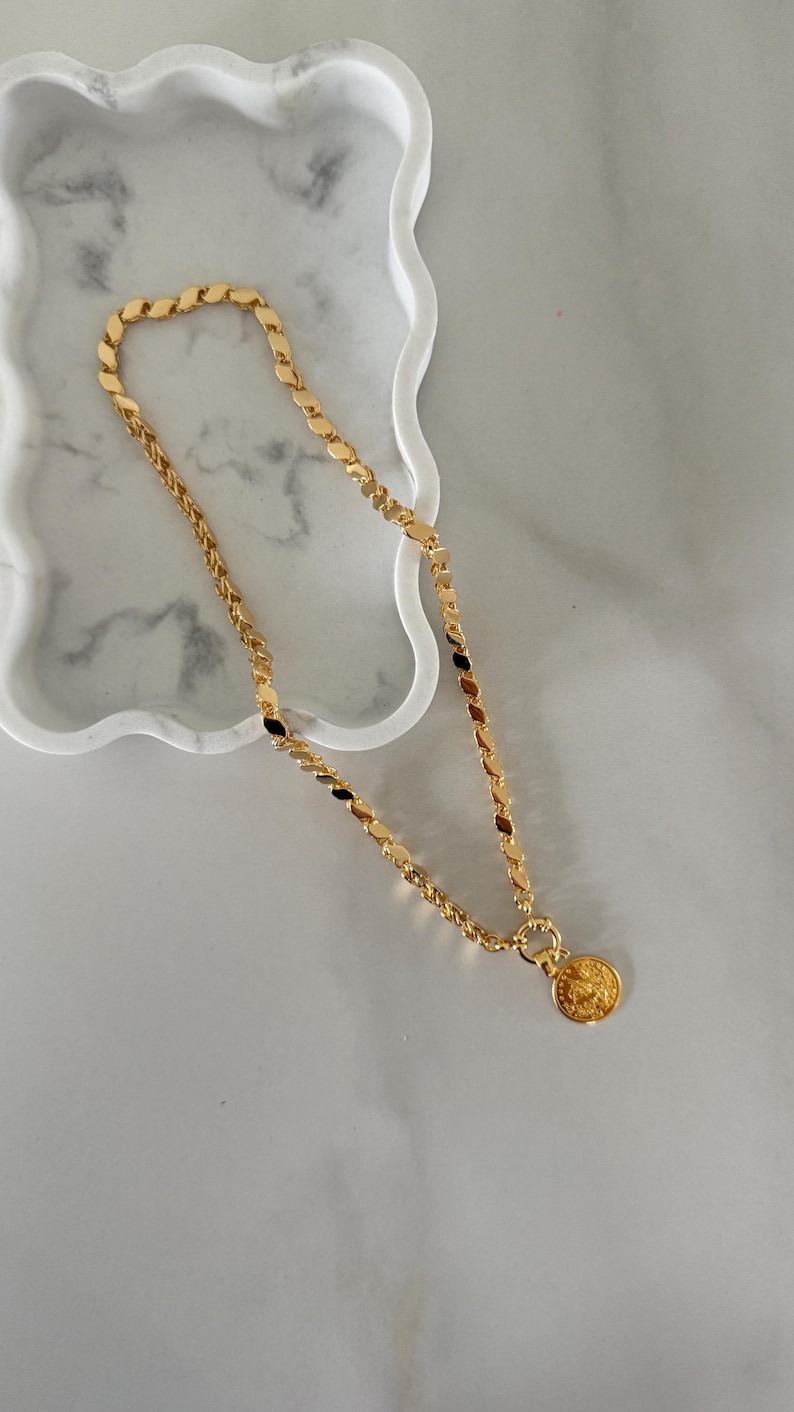 May include: A gold-colored necklace with a coin pendant rests on a white marble tray. The necklace features a variety of chain styles, including a flat, oval link design and a twisted rope chain. The coin pendant has a detailed embossed design.