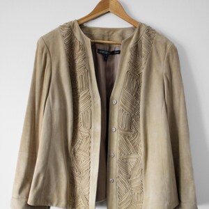 May include: A tan suede jacket with decorative stitched panels down the front and around the collar. The jacket has a button closure and is hanging on a wooden hanger. The brand name "LAFAYETTE 148" is visible on a label.