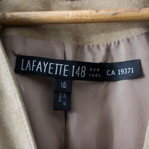 May include: Close-up of a tan suede jacket with a label that reads "LAFAYETTE 148 NEW YORK CA 19371". The jacket is hanging on a wooden hanger. The label also indicates size 16.